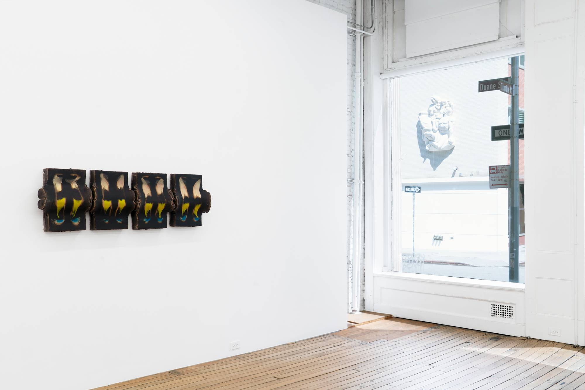 Four nearly identical vertical cuboid ceramic sculptures hang in a tight horizontal line on a white wall in the corner of an art gallery with wood floors. A modern, stone wall-relief sculpture of a family on the facade of a building across the street from the gallery is visible through a large window on the wall adjacent to the four piece sculpture.