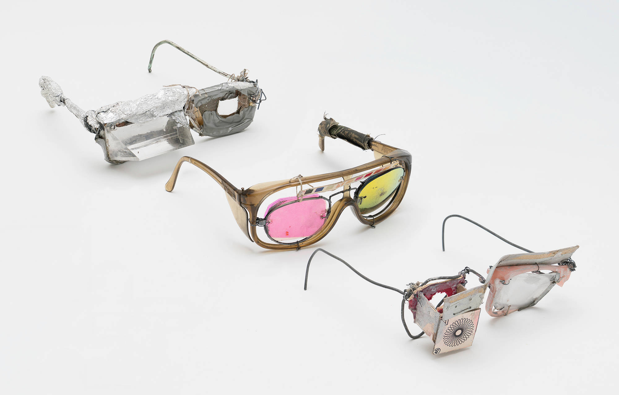 A procession of sculptures resembling homemade glasses constructed out of numerous materials including colored plastics, aluminum foil, and wire.