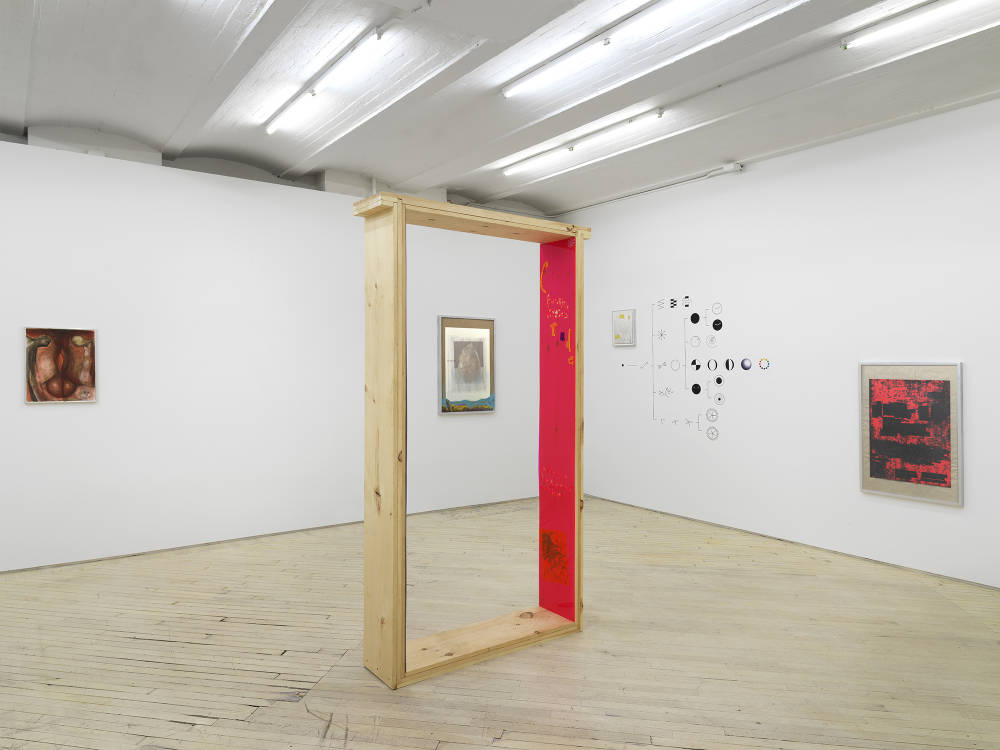 In the center of a gallery space a large wooden rectangular sculpture with one bright pink plastic side. Behind the sculpture is a framed photograph. To its left an abstract painting. To the right a vinyl installation directly on the wall, to its right a framed red and black image. 