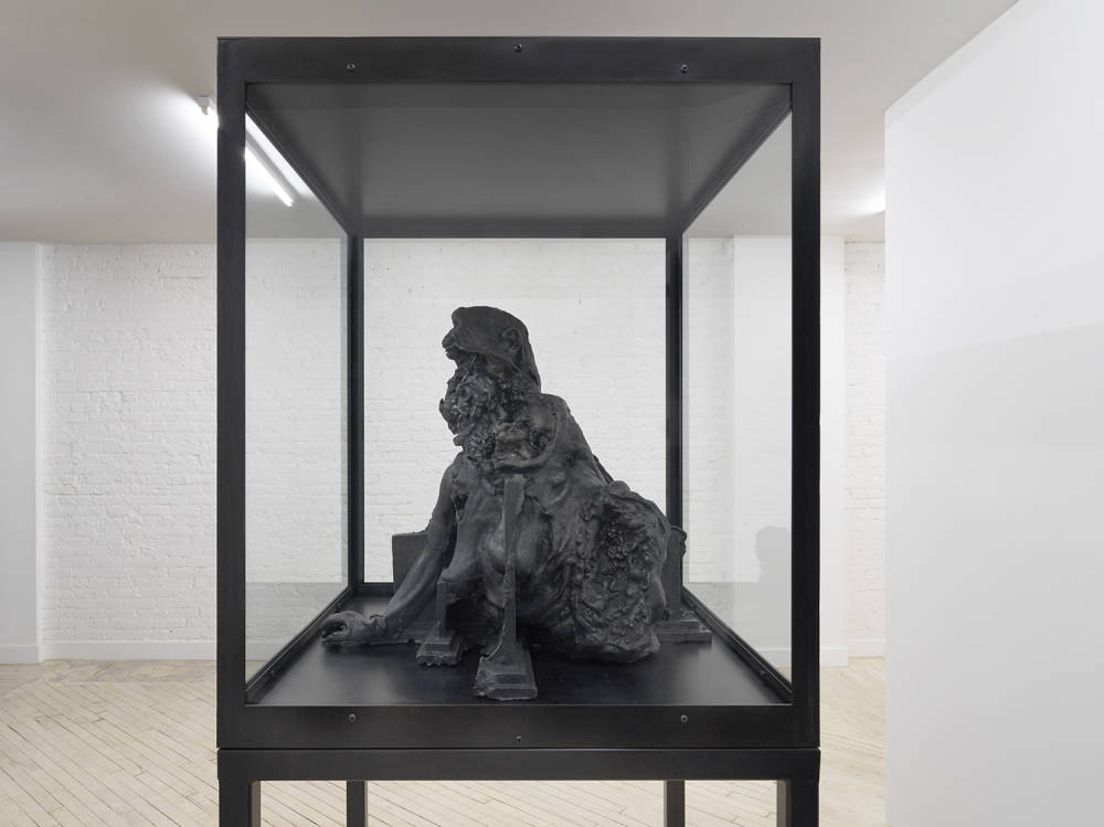 In a gallery space, a large metal and glass vitrine contains a cast iron sculpture resembling a decaying bust form. 