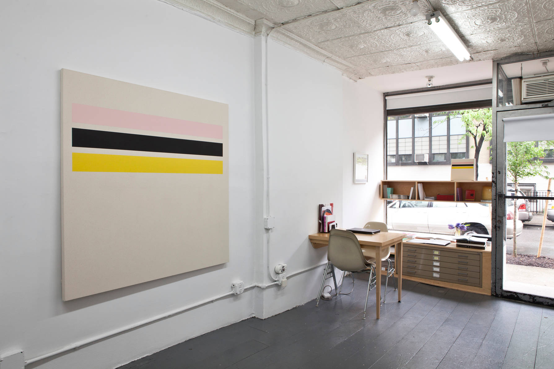 In a gallery space, a large abstract painting is hung on a white wall next to a small desk with two chairs.