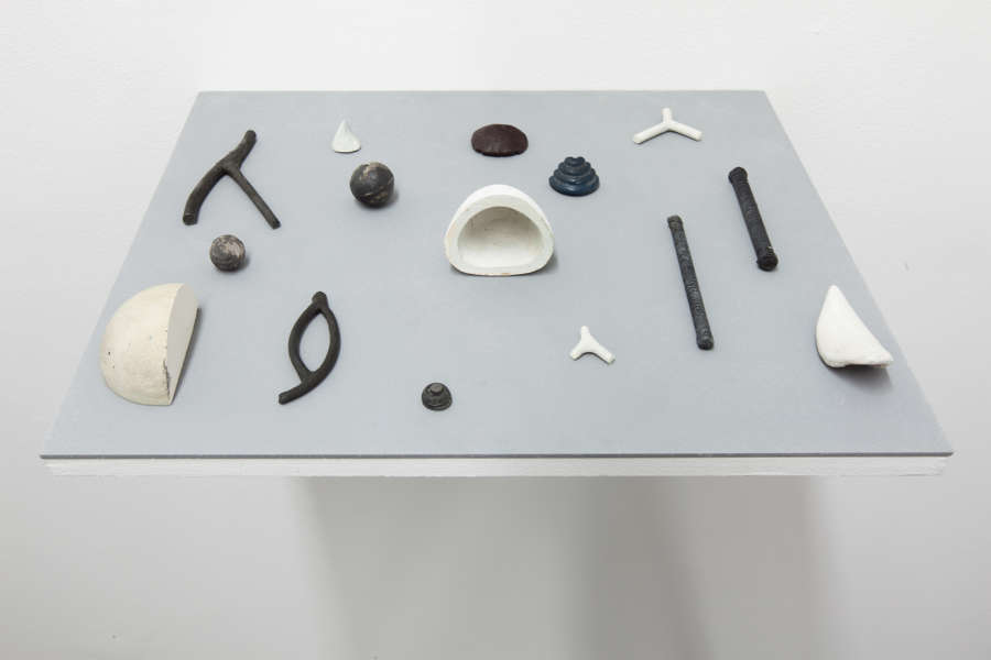 On blue-tinted shelf attached to a white wall, various miniature abstract sculptures and unevenly spaced apart. The objects resemble tools, shell or bone forms 