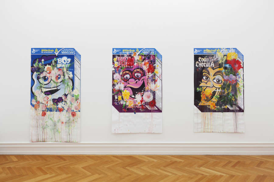 In a gallery space, three abstract paintings are hung in the line depicting printed cereal boxes with numerous abstract brush marks painted on the surface. 