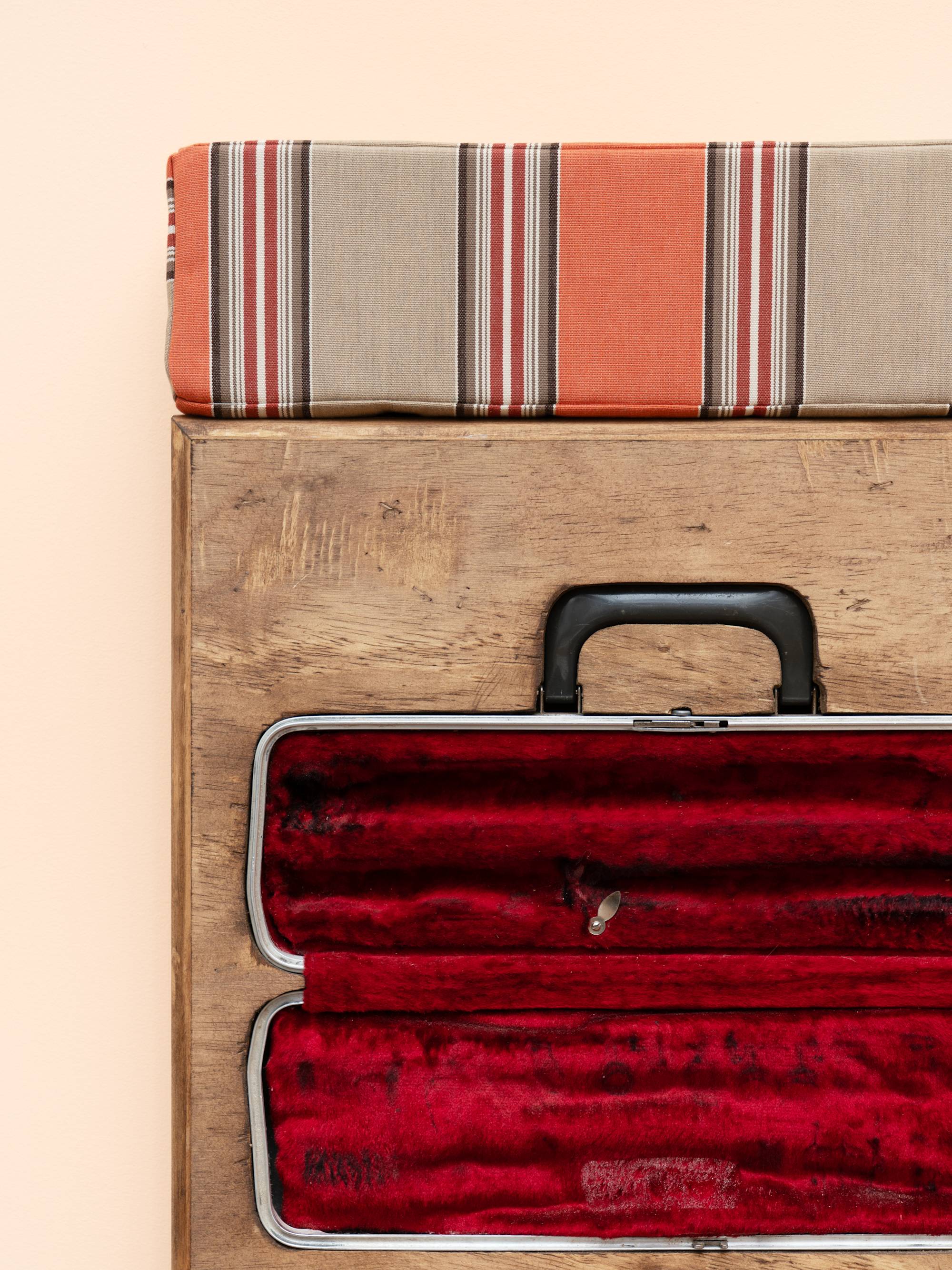 Detail view of a wooden box sculpture with inset flute case with red velvet lining, topped with a orange and beige striped cushion, hanging on a peach colored wall.