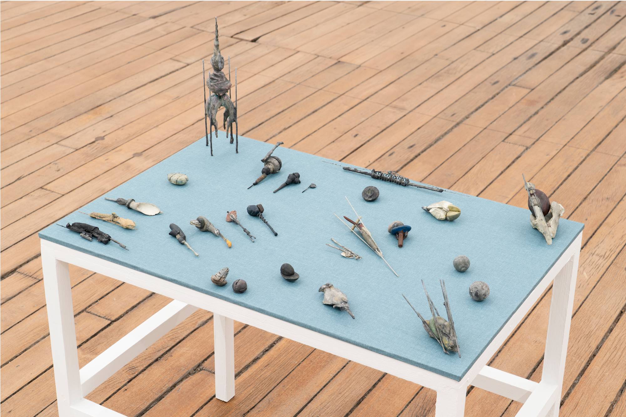 A small, wood table, painted white, with blue fabric top holds an array of abstract miniature sculptures