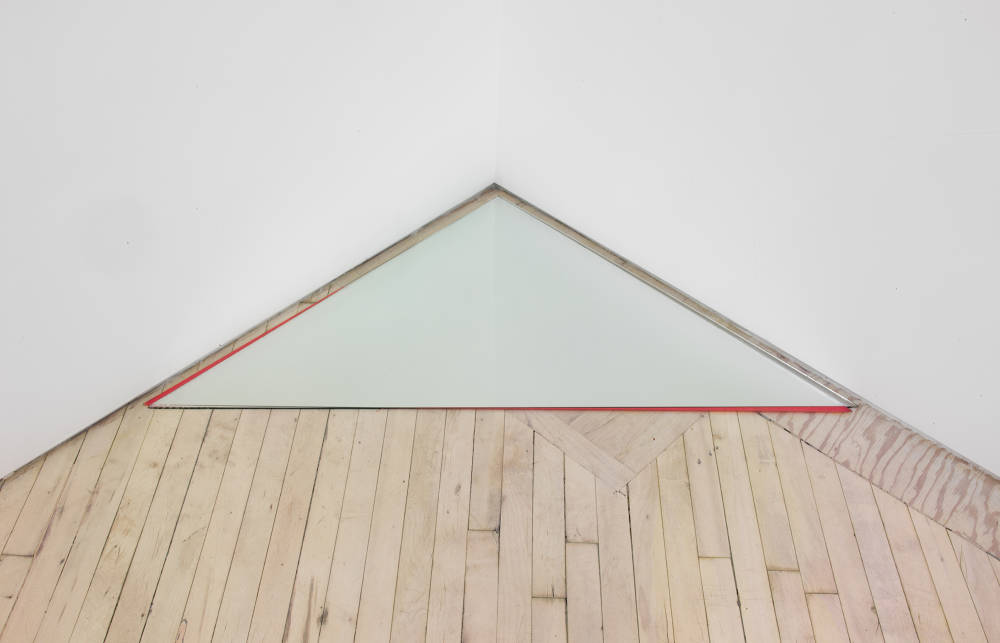 In the corner of a gallery space, a triangular, mirroring object. The object fits precisely within the corner of the room, nearly touching both walls, placed on the floor. 