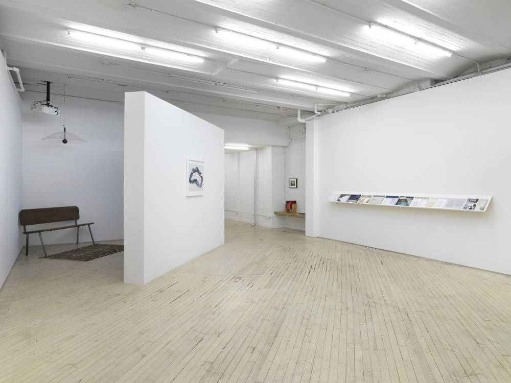In the corner of a gallery space is a darkened projection with a bench. On the front wall a framed artwork. To the left a long shelf holding works on paper. 
