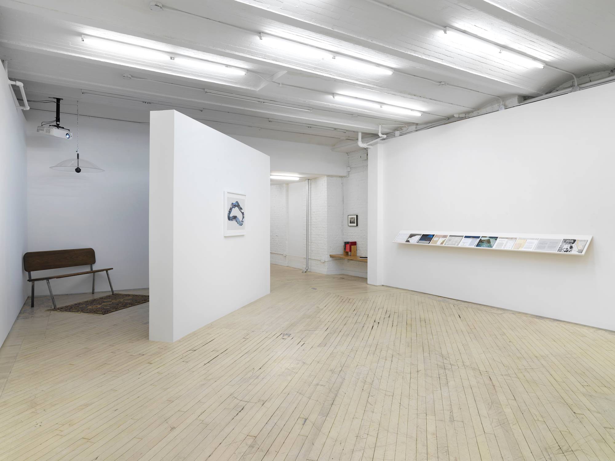 In the corner of a gallery space is a darkened projection with a bench. On the front wall a framed artwork. To the left a long shelf holding works on paper.
