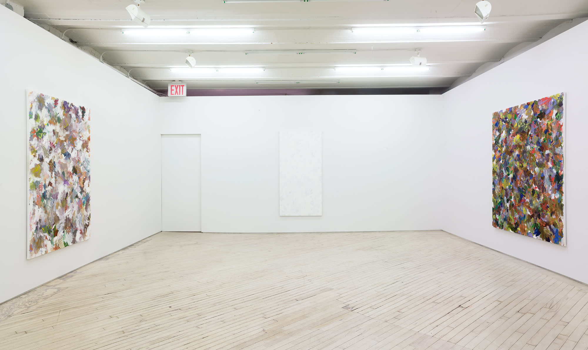 In a gallery space, three large abstract paintings are hung on separate walls. On the center wall there is a doorway to the left with an exit sign above. The paintings contain numerous swatches of various colors painted on top of one another. The paint is thin and in some areas dripping.