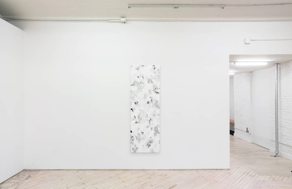 In a gallery space, a large abstract paintings is hung in the center of a large white wall. The painting contains numerous swatches of various colors painted on top of one another. The paint is thin and in some areas dripping. To the right is a hallway leading into another gallery space.