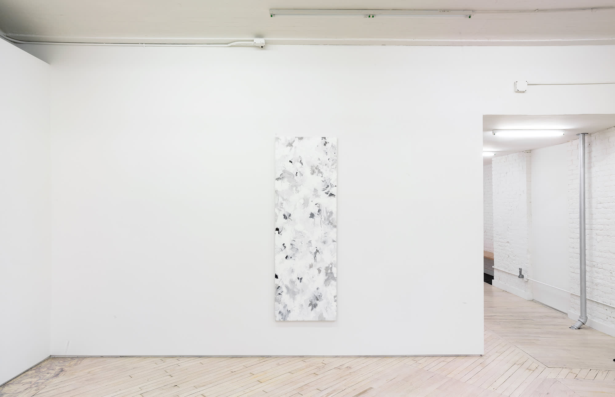 In a gallery space, a large abstract paintings is hung in the center of a large white wall. The painting contains numerous swatches of various colors painted on top of one another. The paint is thin and in some areas dripping. To the right is a hallway leading into another gallery space.