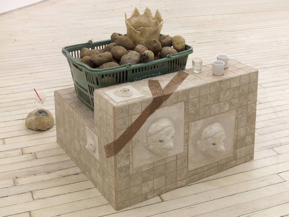 In a gallery, a floor sculpture constructed out of several materials. The base of the sculpture is made out of floor tiles. On its side are multiple reliefs of heads. There are several shot glasses resting on the top of the base. There is a shopping basket full of potatoes. Inside the basket is a crown form containing another relief.