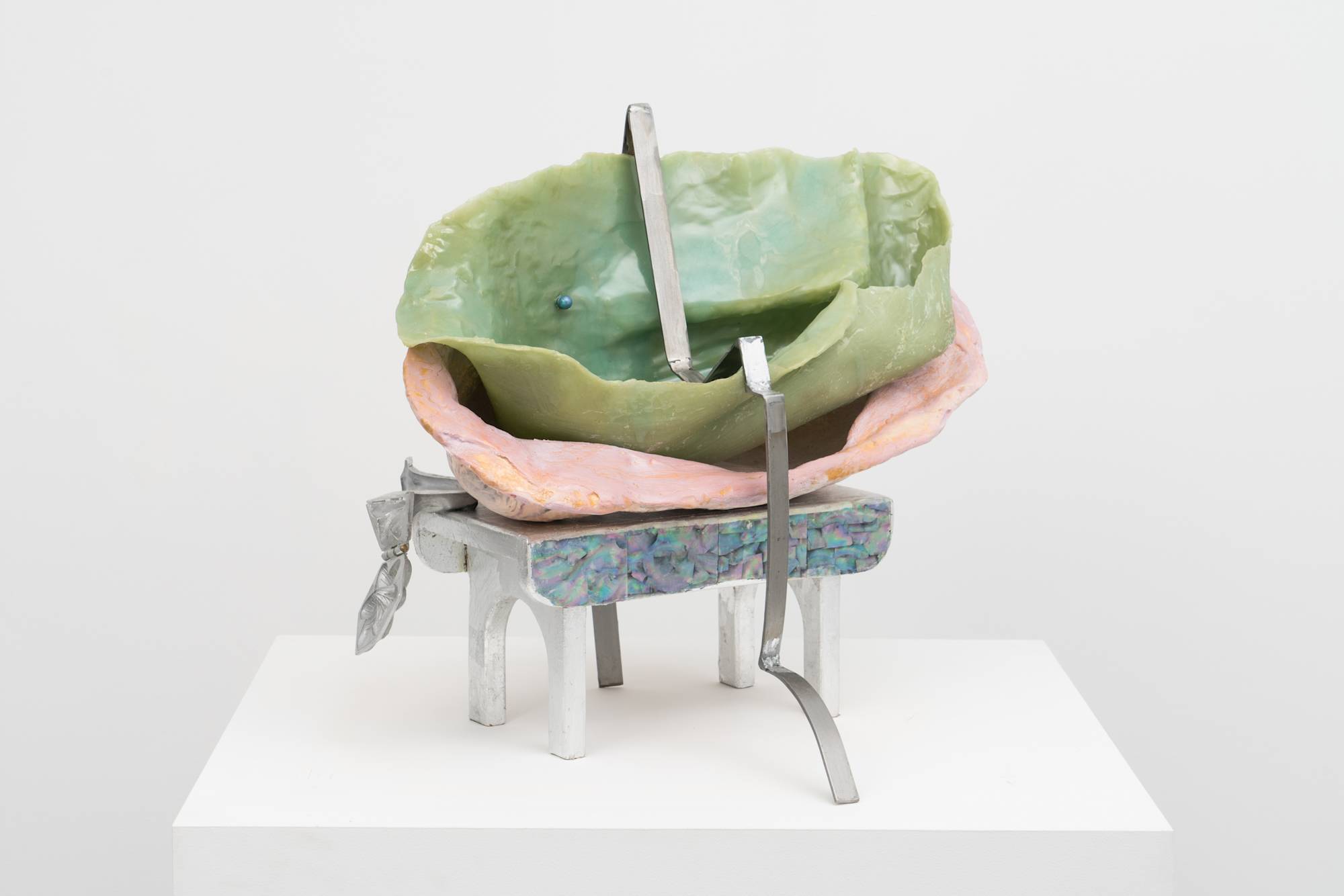 Front view of an Em Rooney sculpture featuring a stool wrapped in foil and mother of pearl, supporting a hinged steel mold of a flower, a pink fiberglass mold of an arm and a thin green silicone cast. A thin strip of bent steel is draped over the sculpture.