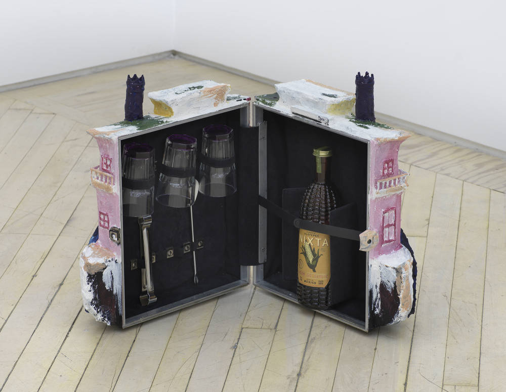 On the floor of a gallery space, a sculpture resembling a boat rendered in pink, yellow, blue, and white. The sculpture is spread open to reveal an inner container that is a traveling bar cart. 