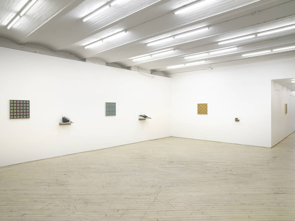 In a large gallery space, three square paintings depicting a grid of crisscrossing lines in various colors. Spaced evenly between the paintings are small, abstract sculptures resembling fossils or other combinations of organic material installed on small gray shelves slightly lower than the paintings. 
