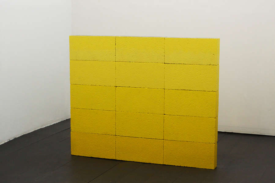 In a gallery space, a stack of concrete slabs painted yellow resembling an anonymous grave. 
