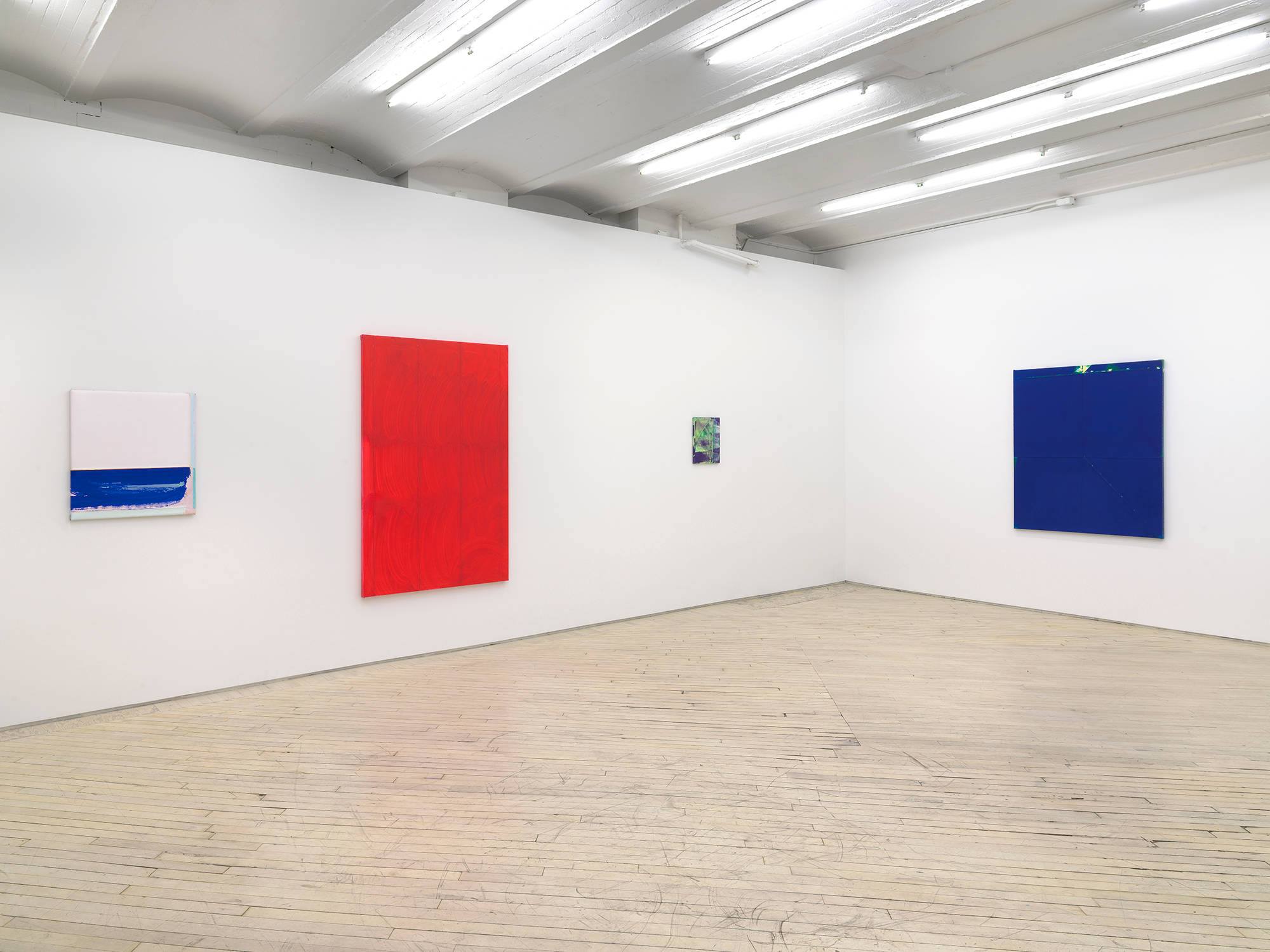Four abstract paintings in a gallery space. To the left a small blue and white square painting. To its right is a larger red monochrome painting with a brushy surface. On the right wall is a large blue monochrome painting. In the center of the larger works is a small brushy green and purple painting.
