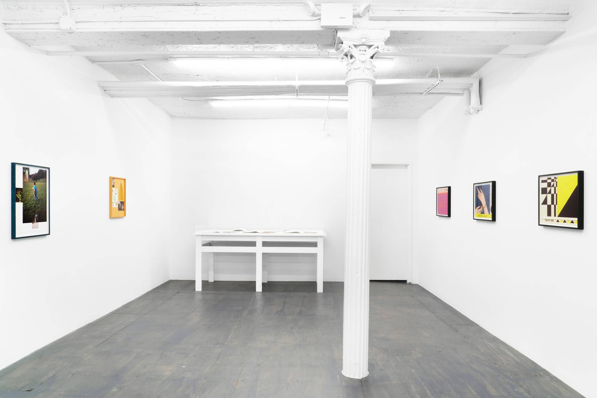 In a gallery space, an assortment of colorful framed photographs are spaced apart with a vitrine placed along the back wall.