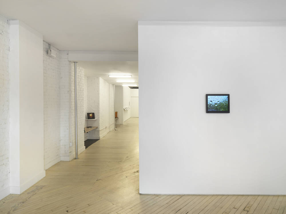 A black digital frame hangs on a gallery wall, to the left is a long hallway with two benches. 