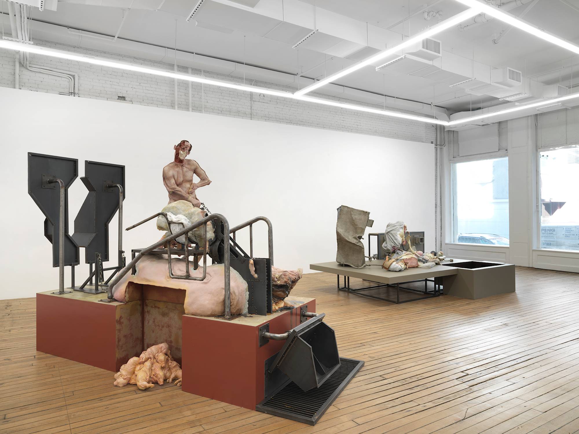 In a large gallery space, two monumental sculptures are spaced apart. Both sculptures contain a combination of abstracted body parts and steel structures raised off the ground.