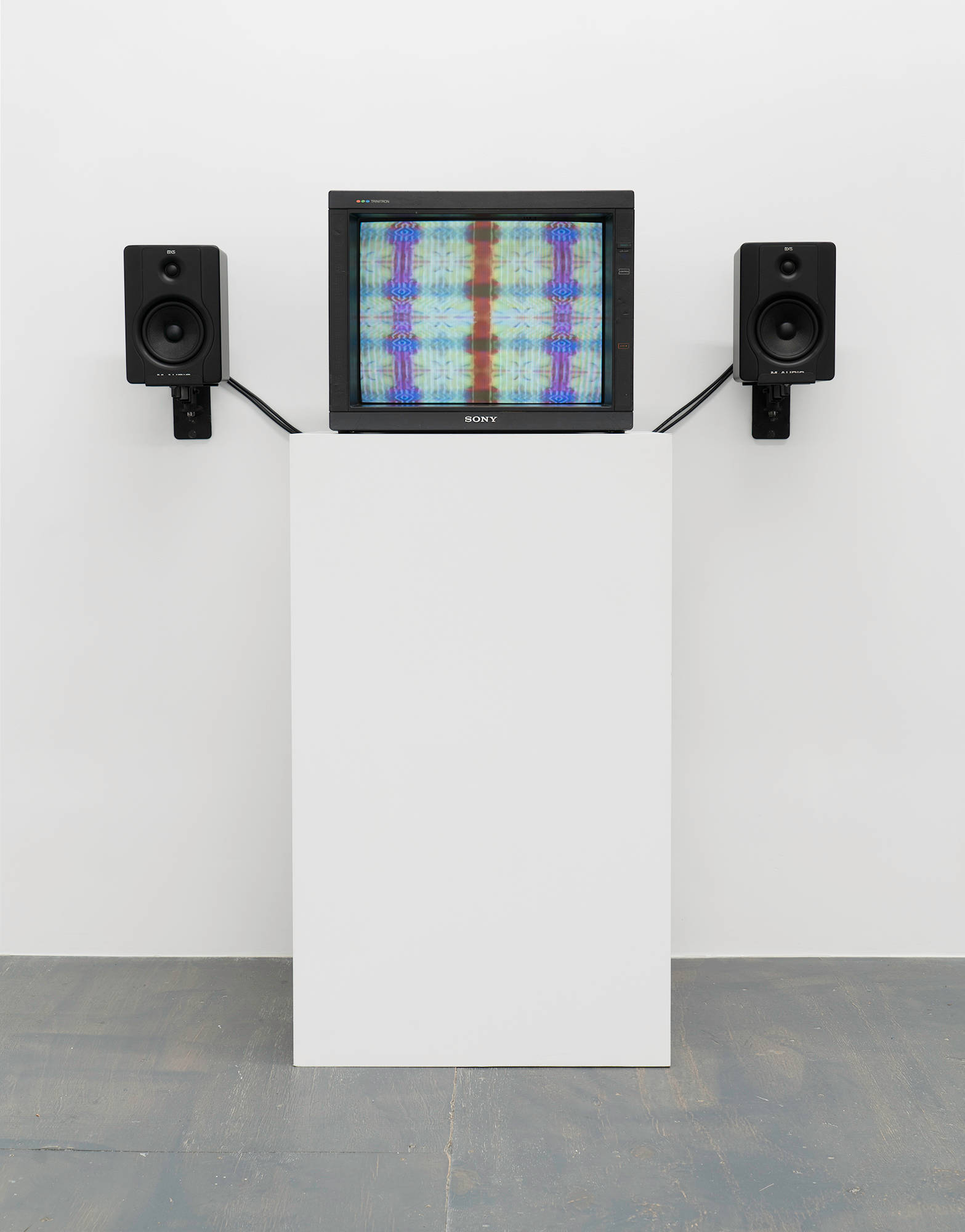 A tv monitor displaying a still image blurred colors in a grid pattern taken from an animation, resting on top of a white pedestal with black speakers mounted to the wall on either side.