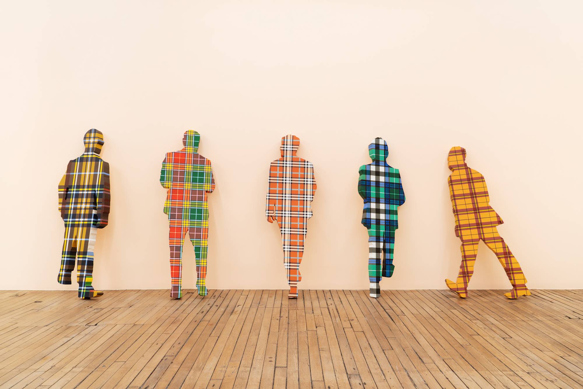 Five flat, life-sized human figures covered in plaid patterns lean against a peach colored wall in an art gallery.