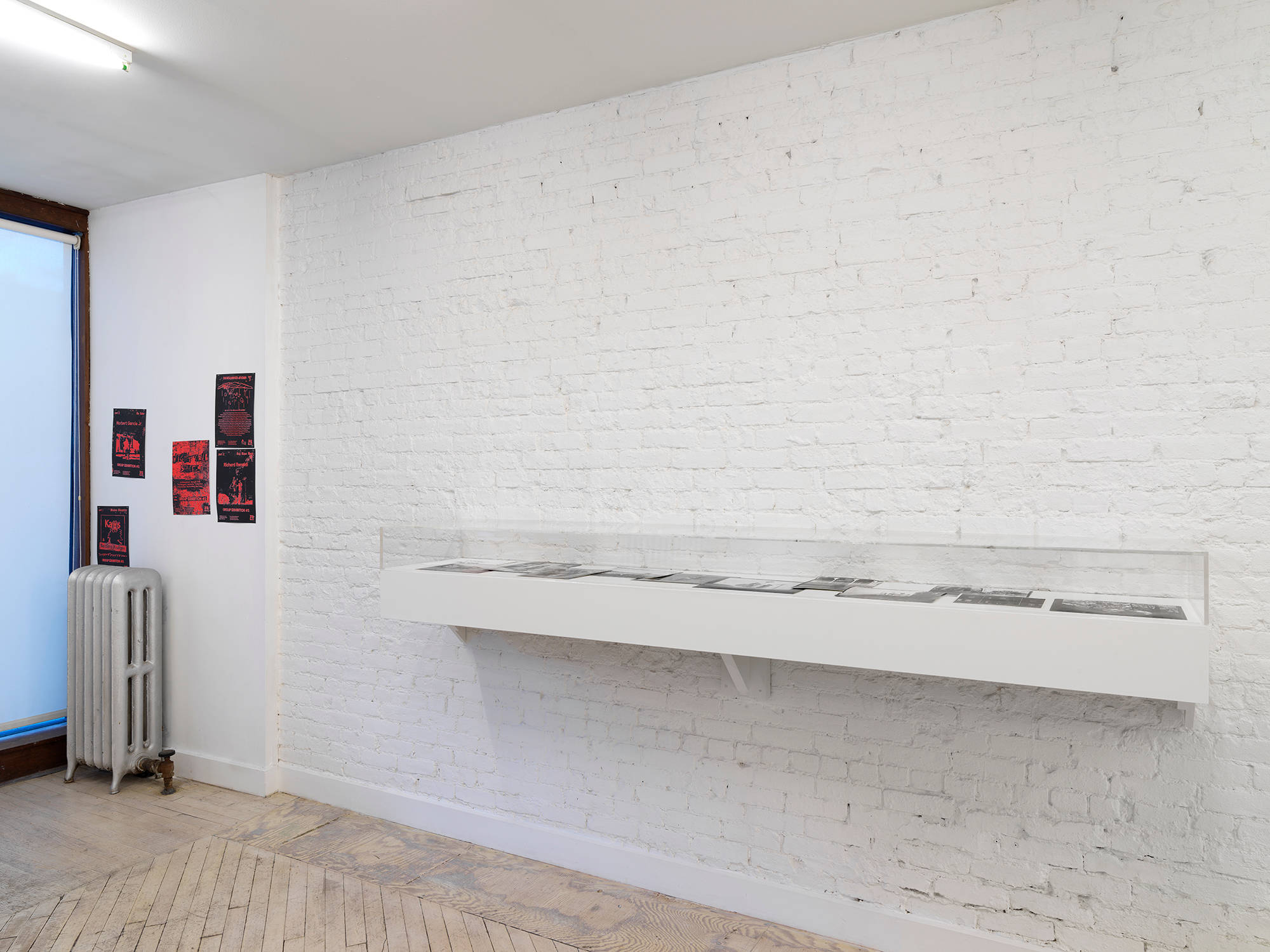 In a gallery space, a wall mounted white vitrine containing black and white photographs, to the left are black and red posters tacked to the wall.