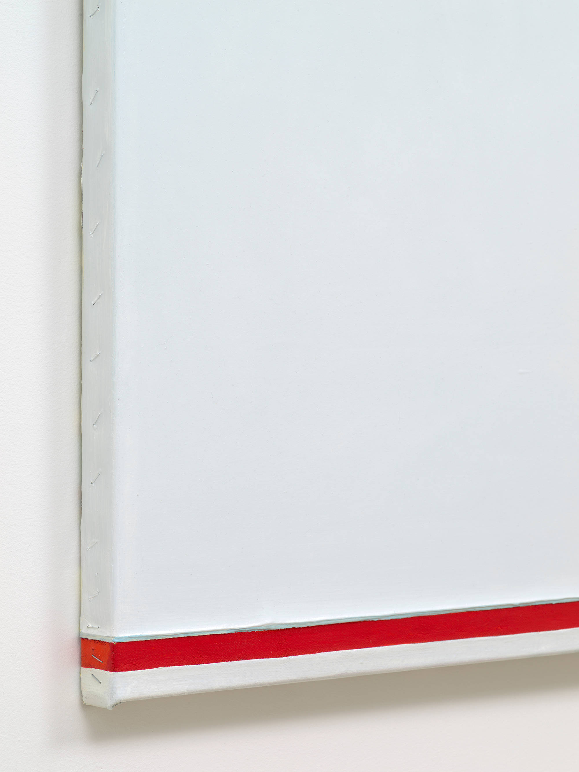 A detail of a white abstract painting. Along the side of the painting is a series of staples. At the bottom edge of the painting is a red bar of color which overlaps onto the side.
