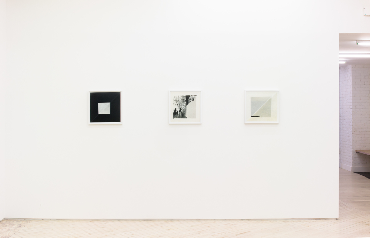 Image of a wall straight on with a view back up the hallway towards the front of the gallery, with 3 small square photos hang on the wall, mostly black and white, and off white of squares of folded book pages with a square format frame.