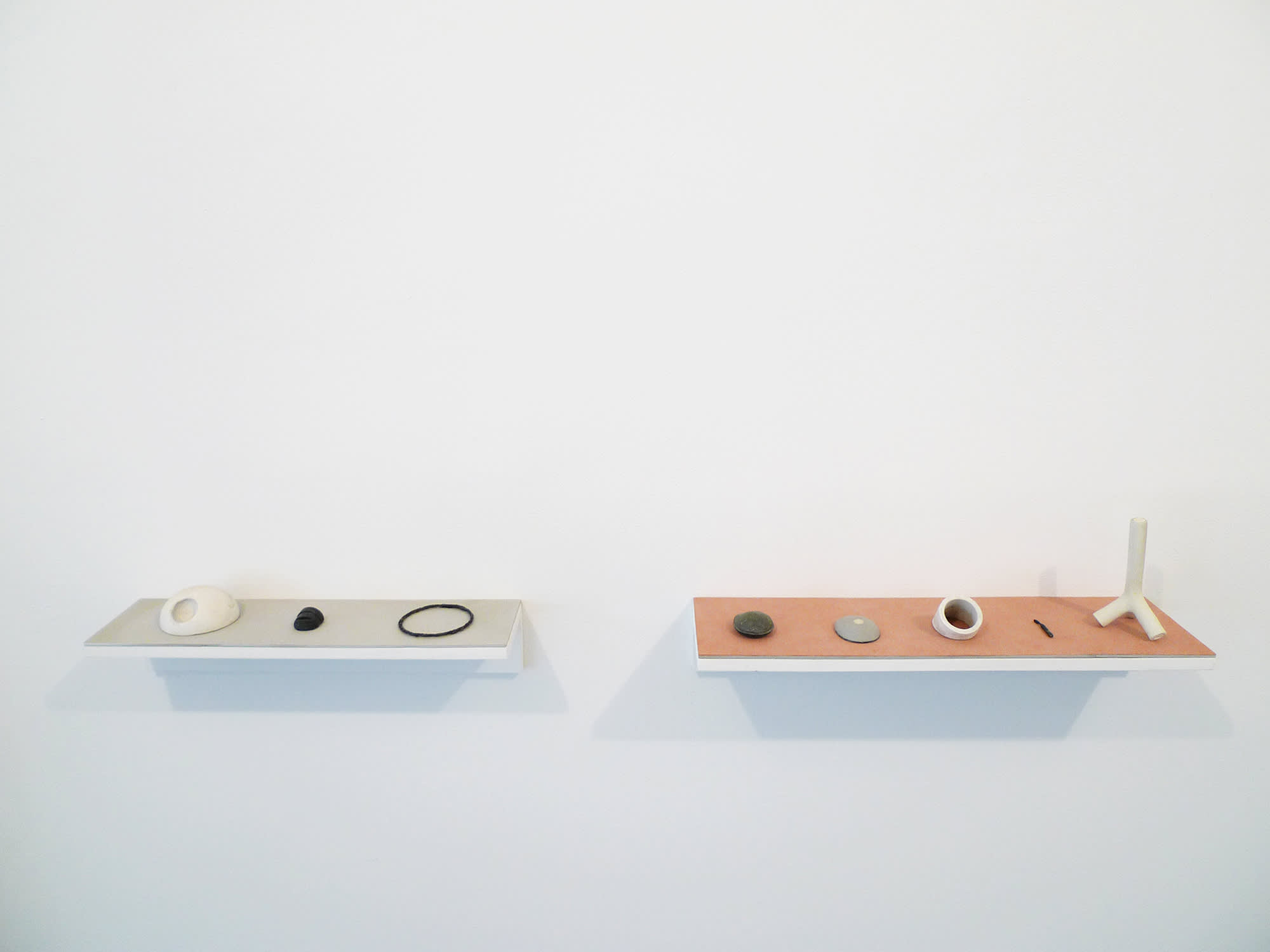 In a gallery space, two shelves are installed in the center of a wall housing numerous handmade objects.