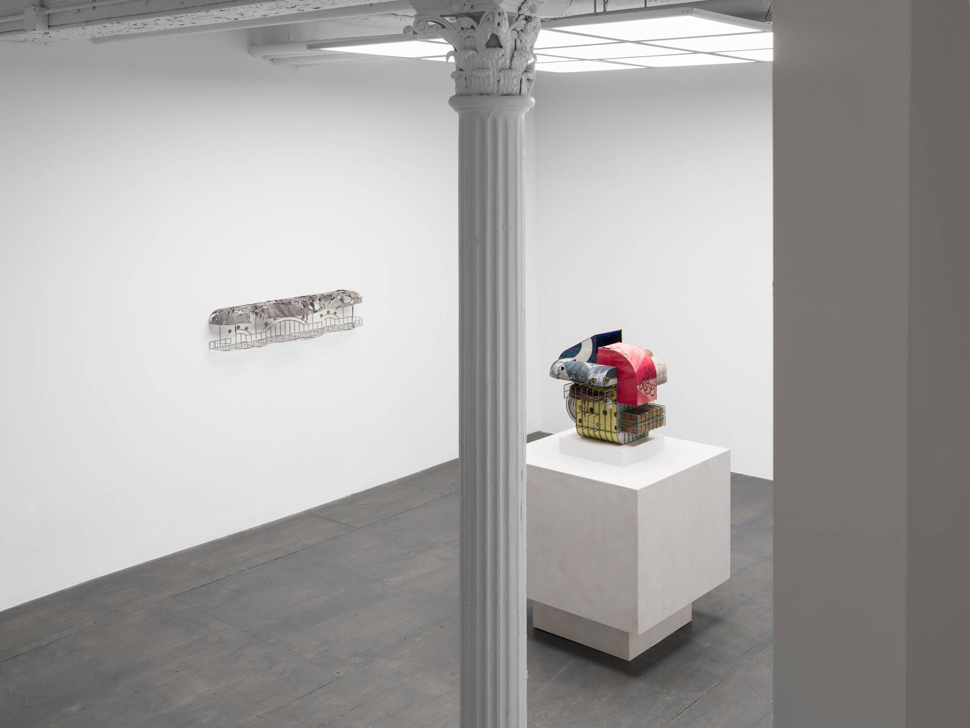 A stairwell view of a white walled gallery with grey floor and white column lit by an LED light fixture that resembles a skylight. Two sculptures are visible in the gallery. One small, silver wall mounted sculpture on the wall to the left and one standing on a pedestal in the center of the room.
