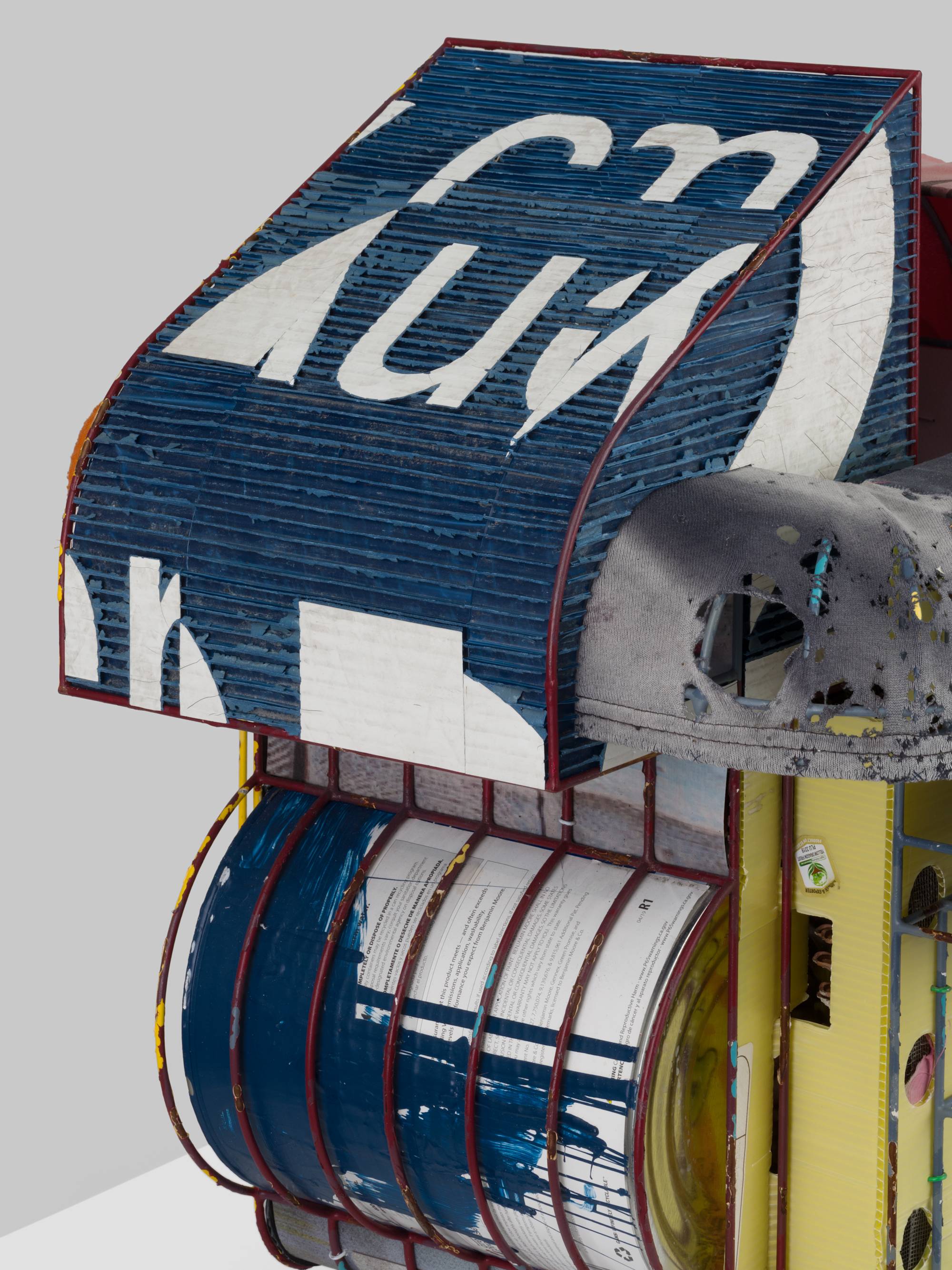 Detail of a cage-like colorful steal wire sculpture, wrapped with several tattered fabrics to resemble various awnings, and embellished with an old shoe, a twig, printed coroplast signage, an old blue paint can, a lock, a paper tag, and a wooden block sits on a white pedestal against a white wall.