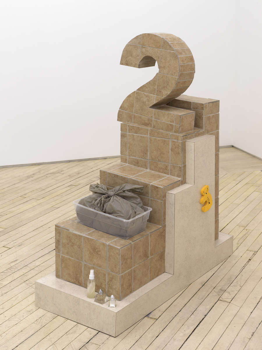 In a gallery space, a large abstract number 2 sculpture constructed out of flooring titles. The base of the sculpture contains two plastic bags and several salt and pepper shakers. 
