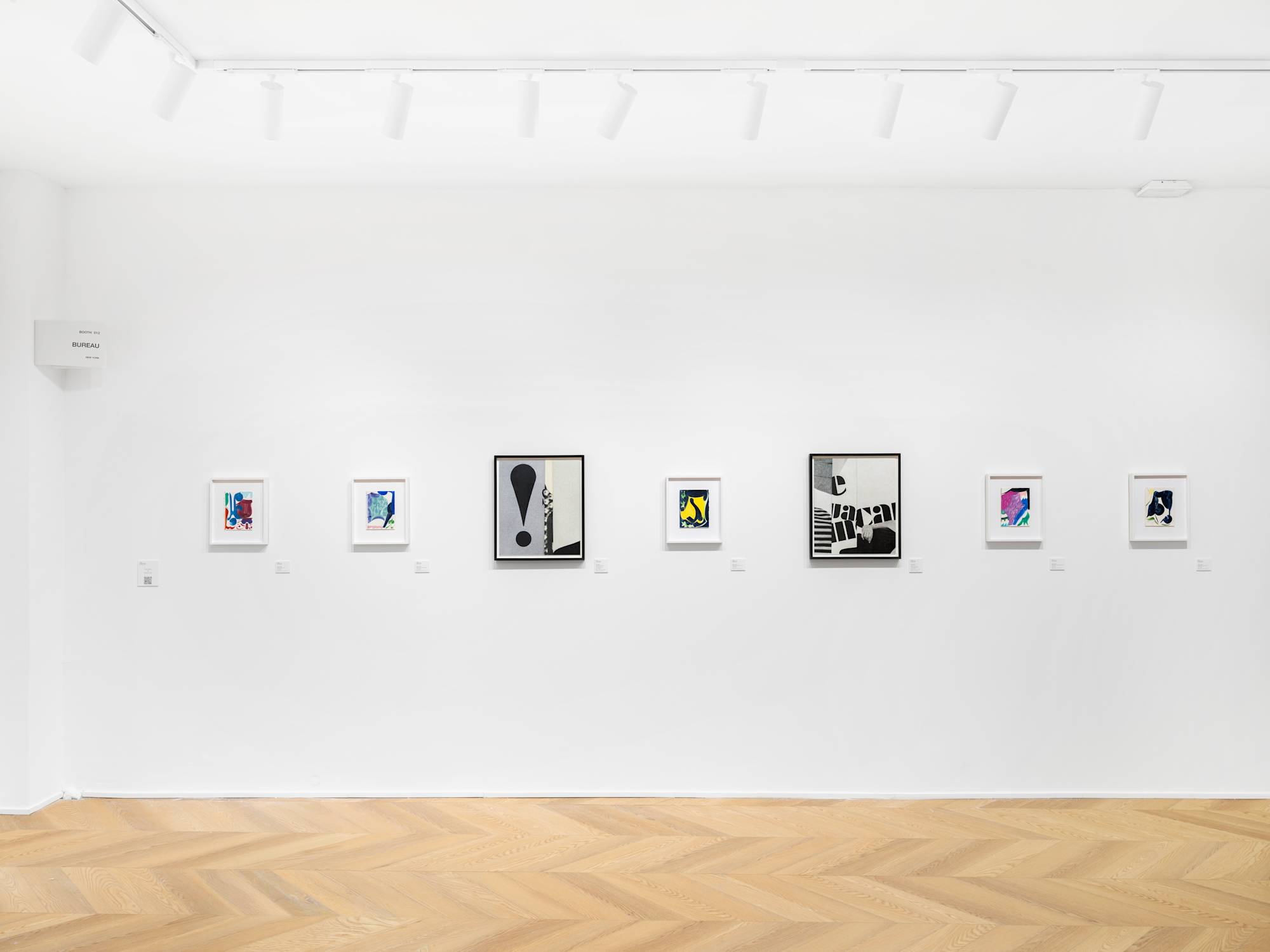 In a gallery space, two black and white framed photographs hang next to small abstract paintings in a range of lush colors.