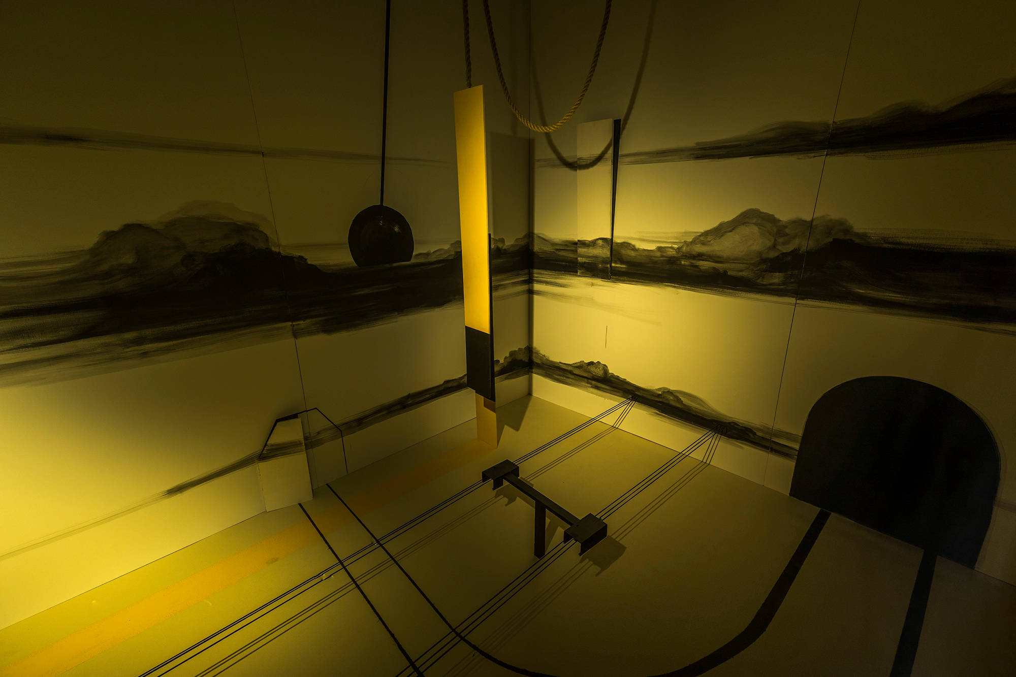 Installation view of a room spotlit with yellow light, a mural on the walls display washed out cloud drawings, a train track is drawn on the floor with sculptures suspended from the ceiling.