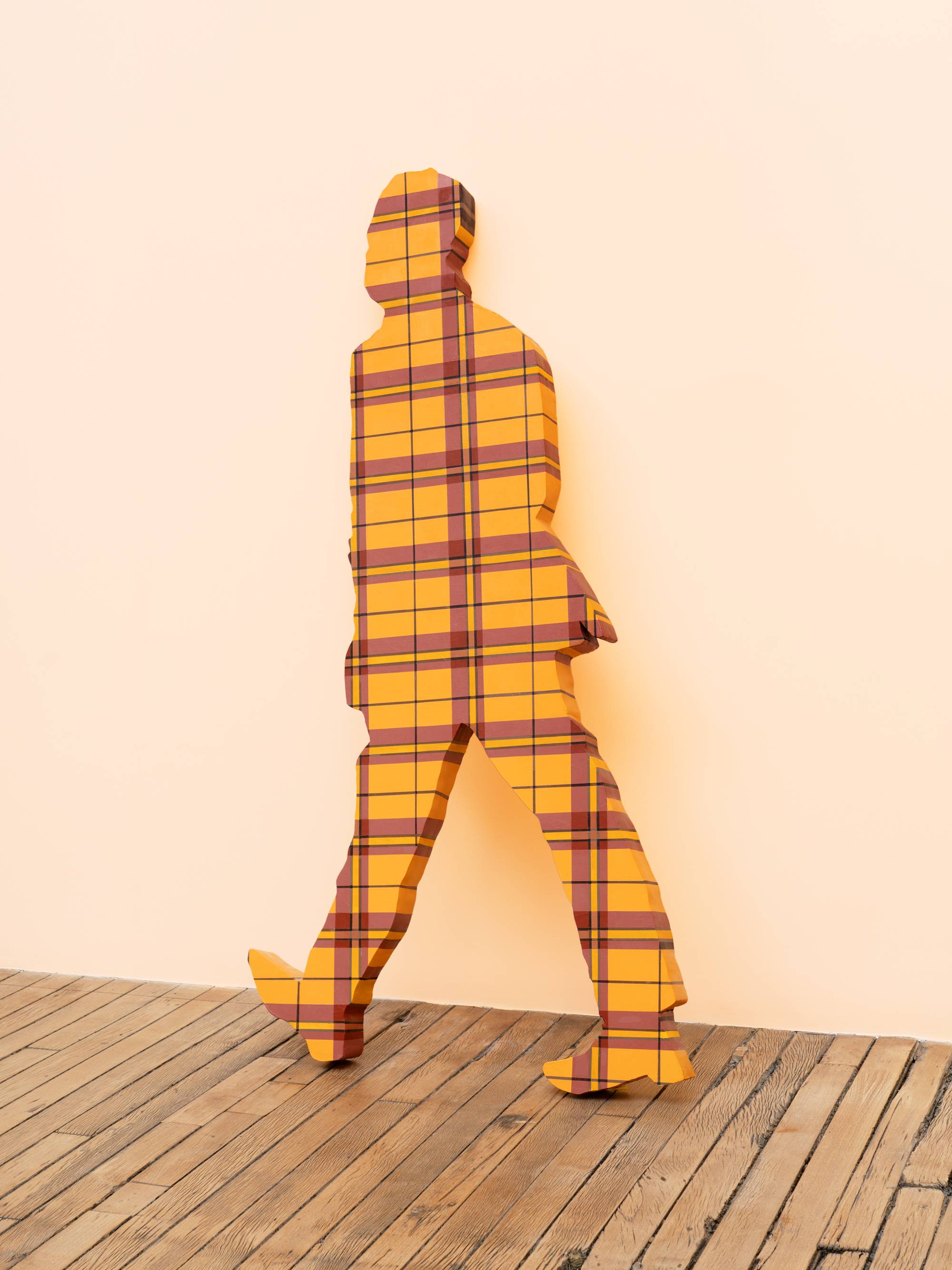 A life-sized human figure painted in a red and yellow plaid pattern leans against a peach wall in an art gallery.