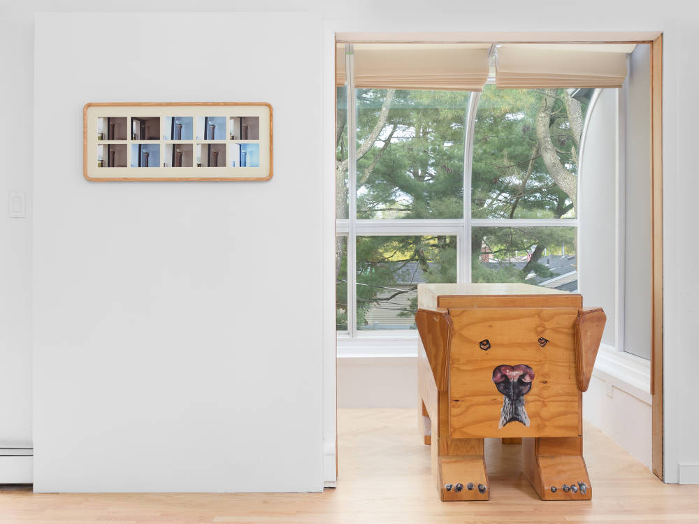 A frame containing multiple small photographs in two rows. To the left a wooden sculpture resembling a dog sits in front of a large window. 