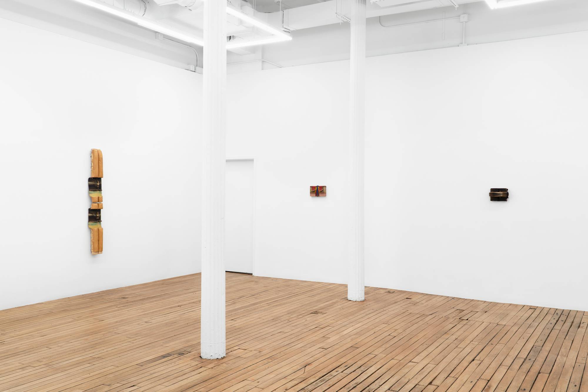 Three wall-mounted clay sculptures of various color and scale hang in the corner of a well lit art gallery with white columns and wooden floor.