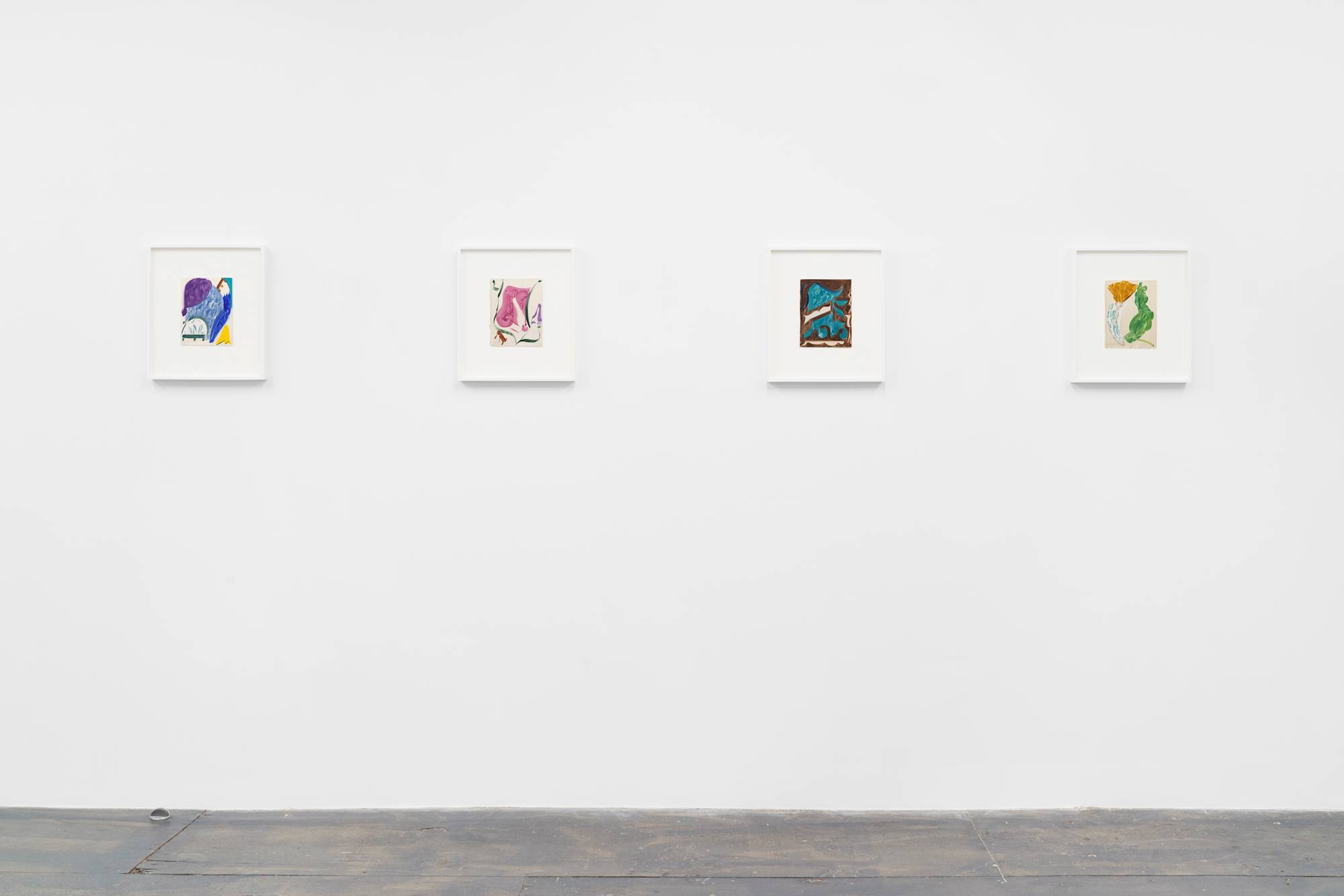Four small, white-framed paintings on paper, of simple shapes and bold colors hang in an art gallery with white walls and grey floor.