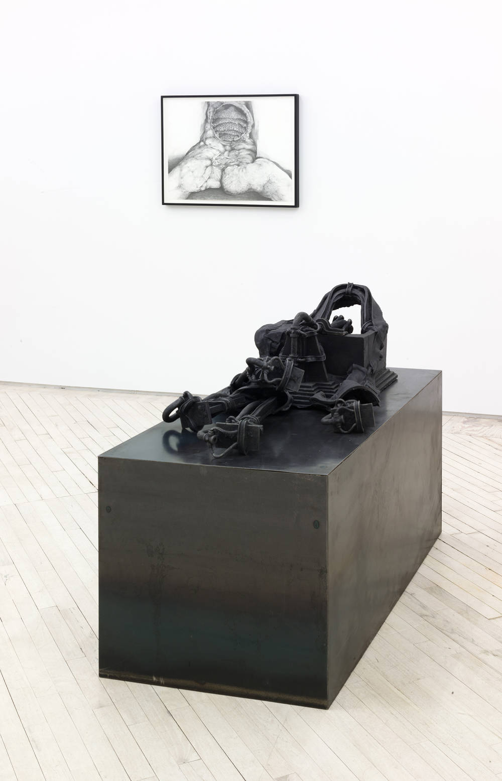 Installation view of a large black sculpture in the foreground and a black and white drawing in the distance. The sculpture has a sleek black metallic, rectangular cube base and on top of it is a complex black object comprised of stirrups or braces and architectural features like low steps and boxes. The drawing at a distance appears to be a mutilated torso, opened up at the abdomen, and perhaps burned or flayed.