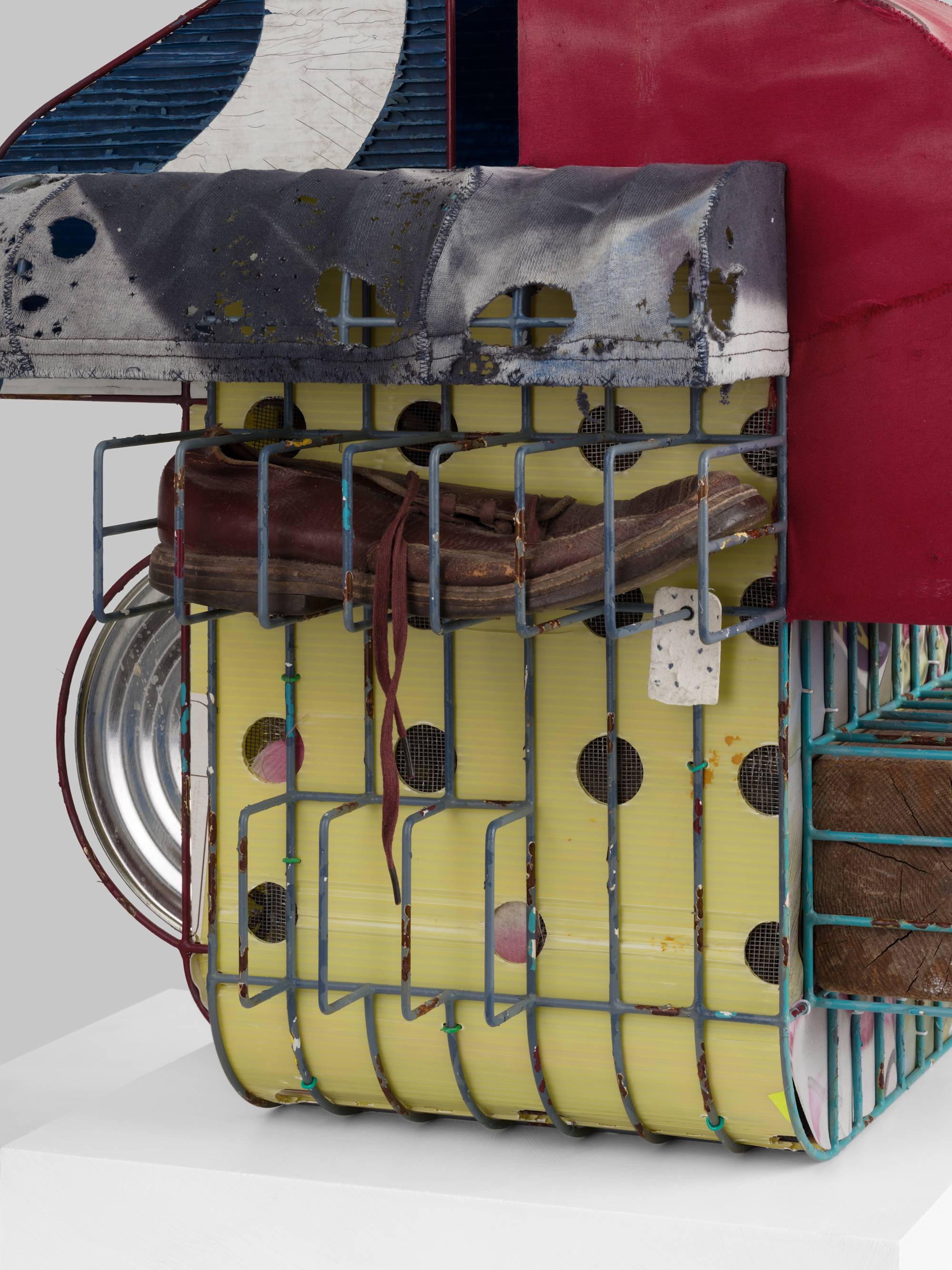 Detail of a cage-like colorful steal wire sculpture, wrapped with several tattered fabrics to resemble various awnings, and embellished with an old shoe, a twig, printed coroplast signage, an old blue paint can, a lock, a paper tag, and a wooden block sits on a white pedestal against a white wall.