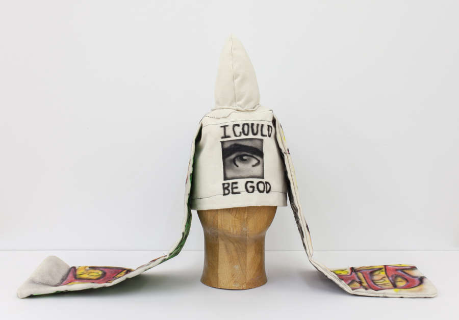 A handmade hat with  long side flaps resting on top of a mannequin. The center of the hat reads "I COULD BE GOD."