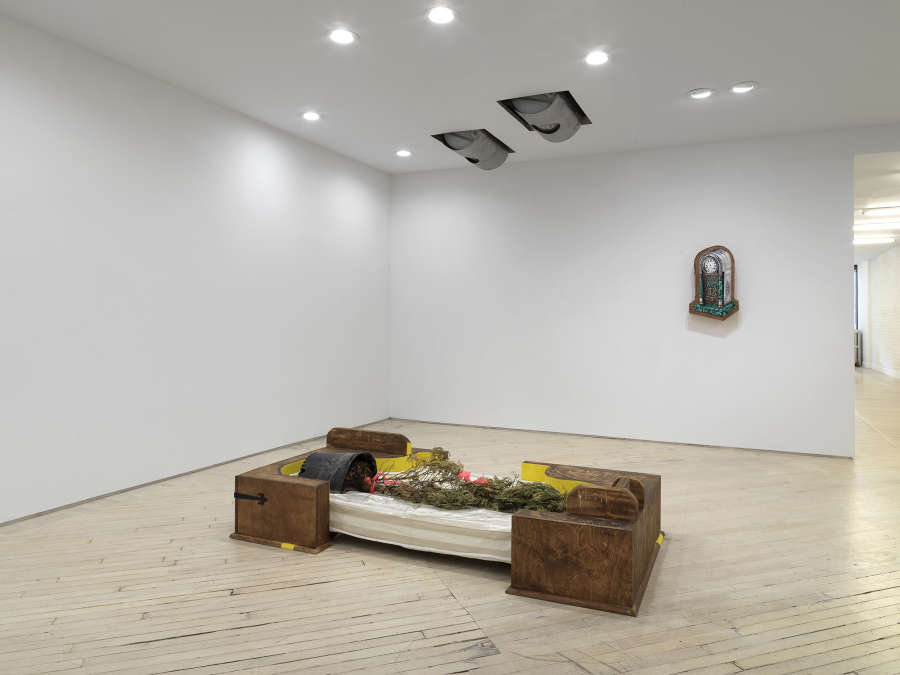 Image of a solo exhibition by Libby Rothfeld showing a wooden sculpture in the foreground and a painting of a clock hanging on the rear wall. There are two metal drums protruding from the ceiling.