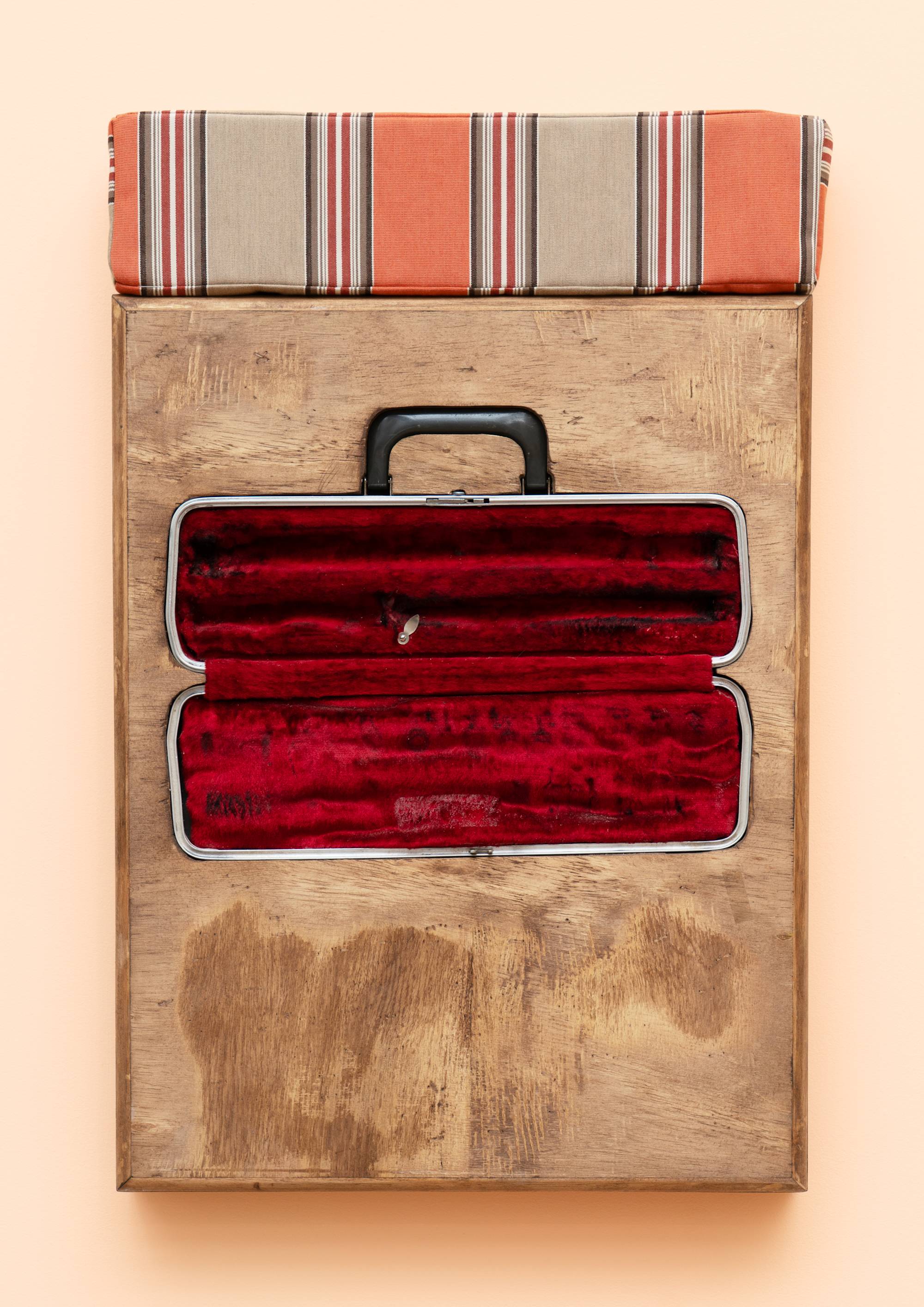 A wooden box sculpture with inset flute case with red velvet lining, topped with a orange and beige striped cushion, hanging on a peach colored wall.
