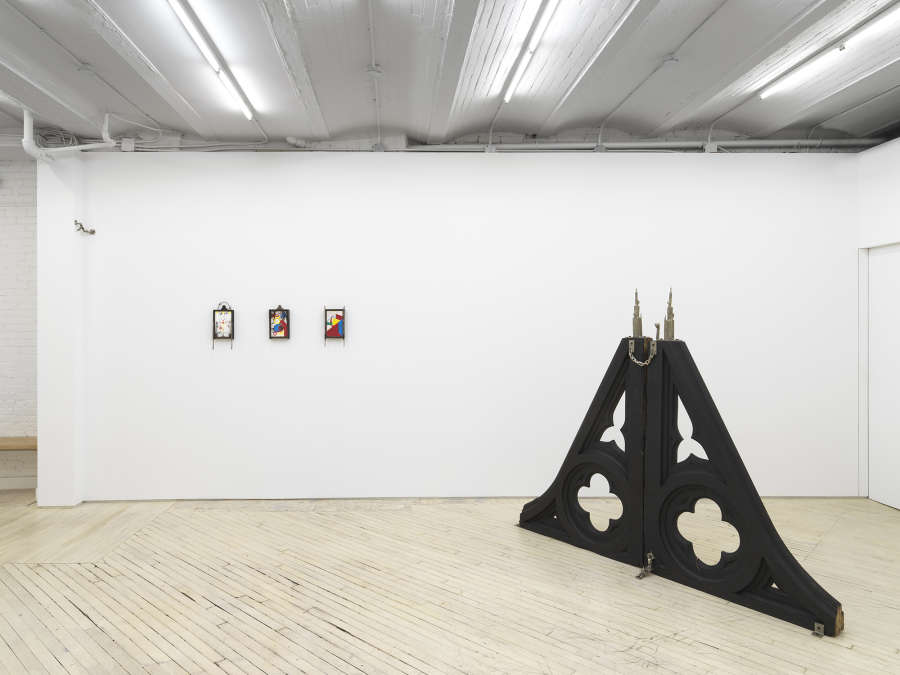Installation view of main gallery: at left three small colorful drawings hang in the wall, framed with ornate black wood. To the right is a pyramid shaped gothic style wooden sculpture which is made of two giant gothic corbels placed together, at the top of the pyramid are 2 pointy cast bronze objects and a cast bronze chain connecting the two.