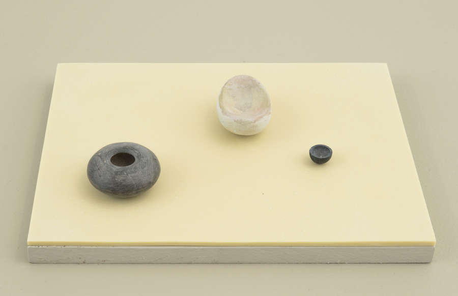 Three miniature sculptures resembling objects found in nature placed on top of a yellow-tinted plinth. 