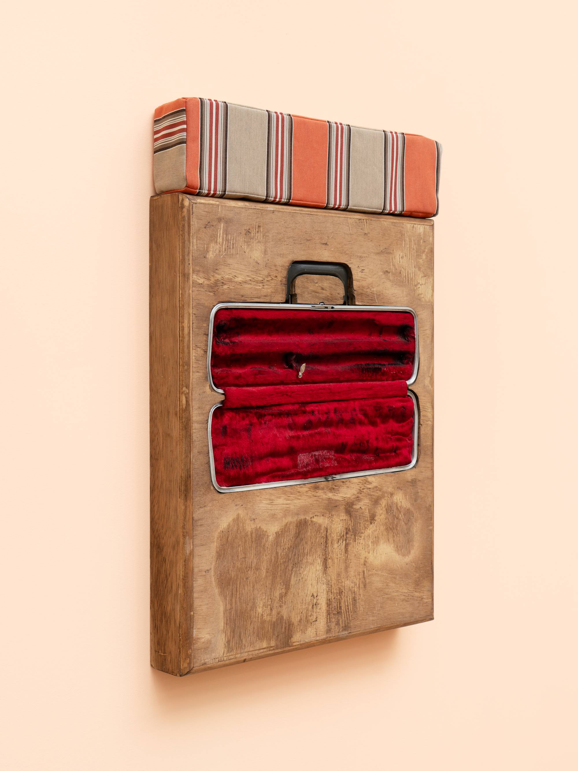 Angle view of a wooden box sculpture with inset flute case with red velvet lining, topped with a orange and beige striped cushion, hanging on a peach colored wall.