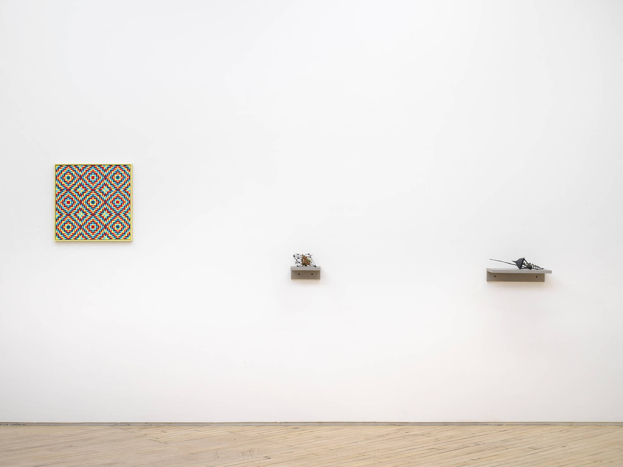 On a long white wall to the left a square painting depicting a grid of crisscrossing lines rendered primarily in red and yellow. To the right two small abstract sculptures resembling handmade artifactual tools installed on small gray shelves.