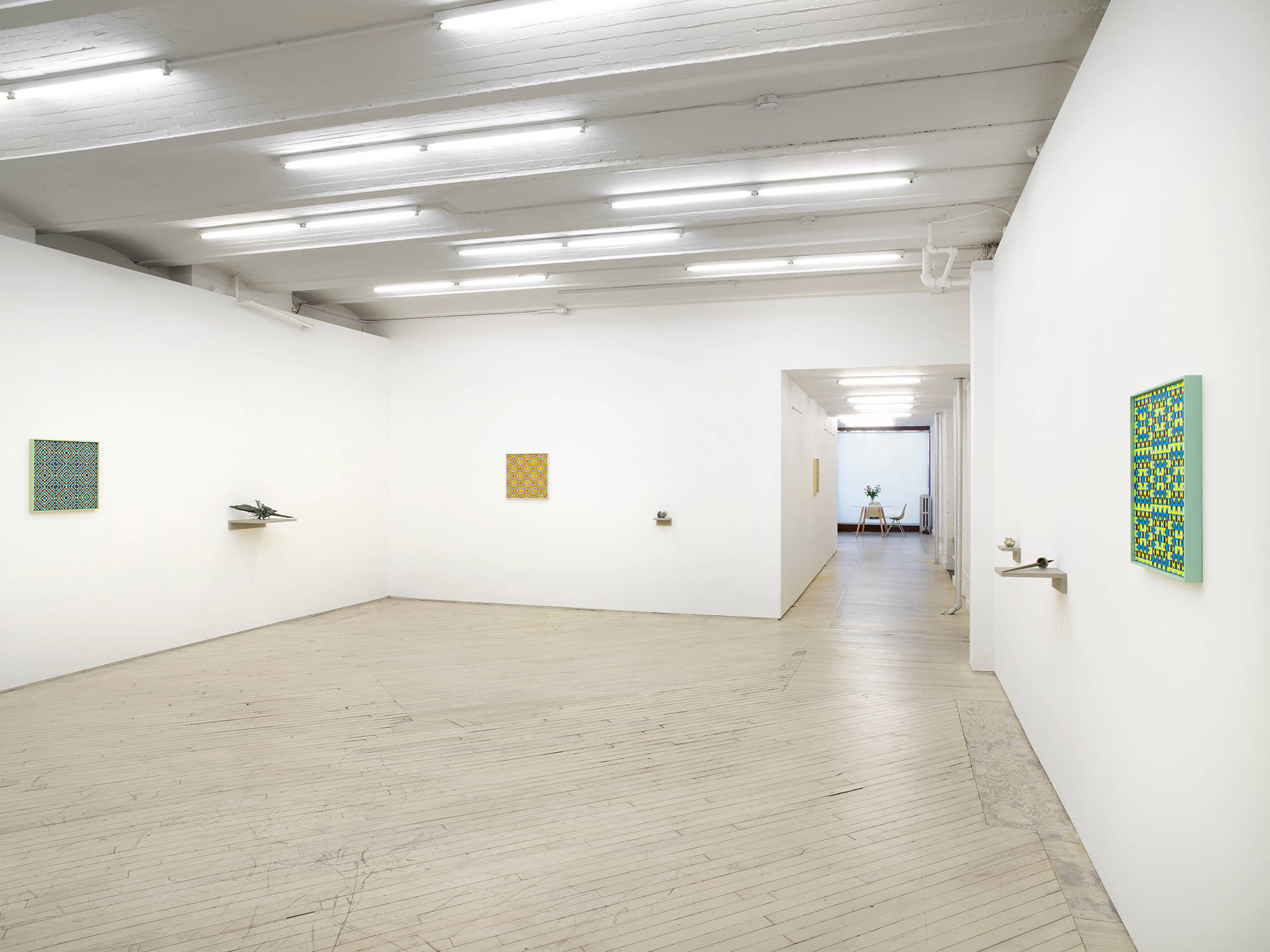 In a large gallery space, three square paintings depicting a grid of crisscrossing lines in various colors. Spaced evenly next to the paintings are small abstract sculptures resembling fossils or other combinations of organic material installed on small gray shelves slightly lower than the paintings. To the right is a hallway leaving to an additional gallery space with a large window.