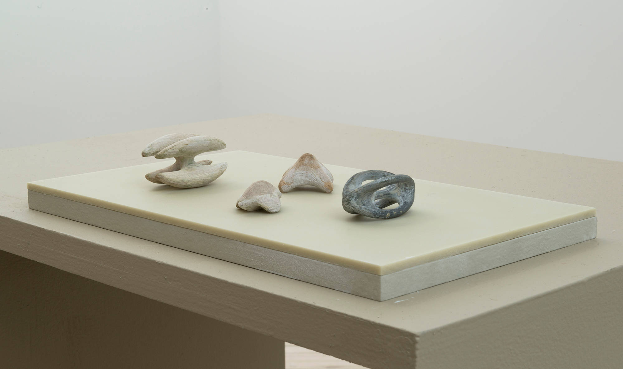 Four miniature sculptures resembling organic forms on top of a green-tinted plinth. The table housing the plinth is visible with a white wall in the background.